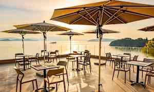 Udendørs servering med borde og parasoller ved havet i solnedgang. (AI-genereret tekst)