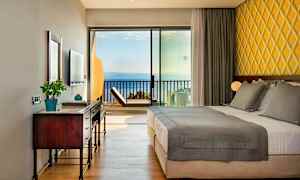 Hotelværelse med dobbeltseng, skrivebord og havudsigt gennem glaspanel. (AI-genereret tekst)