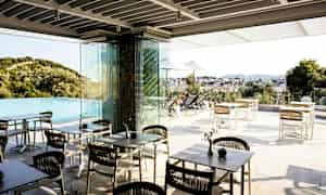 En udendørs terrasse med borde og stole nær en infinitypool med udsigt over et landskab og en by. (AI-genereret tekst)