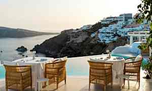 Udendørs område med borde og stole ved poolen, med udsigt over kysten og hvide bygninger på en bakke. (AI-genereret tekst)