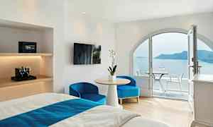 Hotelværelse med lyse hvide vægge, møbleret med en seng, bord og stole, og en balkon med udsigt over havet og en ø. (AI-genereret tekst)