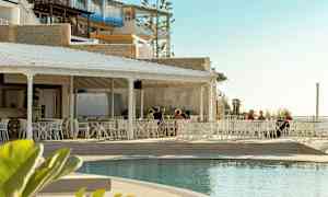 En udendørs restaurant med hvide møbler ved siden af en pool, med nogle personer der sidder ved bordene. Baggrunden består af bygninger og en lys himmel. (AI-genereret tekst)