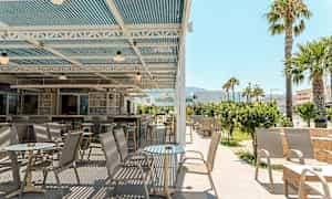 En udendørs terrasse med borde og stole under et overdækket tag, omgivet af grønne områder og palmer. (AI-genereret tekst)