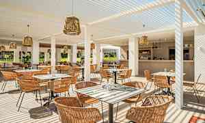 En udendørs restaurant med siddepladser og borde, dækket med hvide pergolatag og pyntet med rottinglamper. Baren er synlig i baggrunden. (AI-genereret tekst)