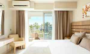 Et hotelværelse med en seng, et skrivebord og en stol samt en balkon med udsigt over palmer. (AI-genereret tekst)