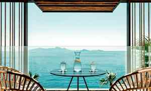 Et udendørsområde med et bord, hvorpå der står en kande og tre glas med vand, med udsigt over et stille hav og fjerne øer. (AI-genereret tekst)