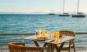 Et bord med morgenmad og juice står ved havet, med sejlbåde i baggrunden. (AI-genereret tekst)