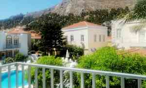 Billedet viser et udendørsområde med swimmingpool, bygninger og et bjergrigt landskab i baggrunden. (AI-genereret tekst)