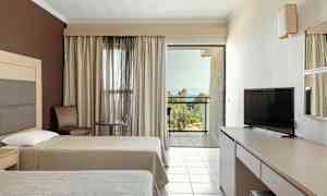 Hotelværelse med to senge, tv, bord og stole samt en balkon med udsigt over grønne områder og hav. (AI-genereret tekst)