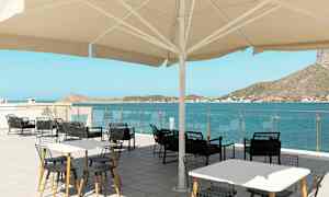 Udendørs serveringsområde med borde og stole under en stor parasol, med udsigt over havet og omkringliggende bakker. (AI-genereret tekst)