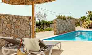 To solsenge med et håndklæde og en stråtaske ved siden af en pool. Der er også en stenmur og palmer i baggrunden. (AI-genereret tekst)