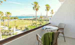 En balkon med bord, stole og udsigt over en grøn have med palmer samt havet i baggrunden. (AI-genereret tekst)