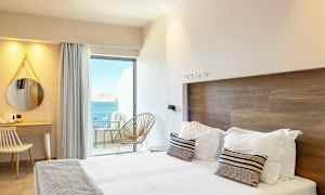 Et hotelværelse med moderne indretning, en dobbeltseng, et skrivebord og udsigt over havet fra en altan. (AI-genereret tekst)