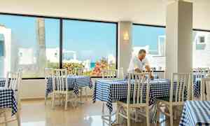 Lys restaurant med hvide stole og ternede blå duge, samt store vinduer med udsigt over bygninger og grønne områder. (AI-genereret tekst)