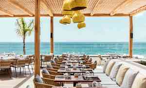 Restaurant med havudsigt, med borde og stole i træ og flet samt bænke med puder under et beskyttende tag. (AI-genereret tekst)