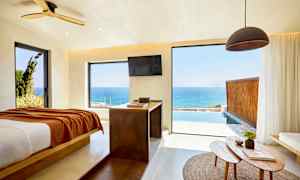 Et rum med udsigt over havet gennem store vinduer, en seng er placeret til venstre og et bord med genstande er i forgrunden. (AI-genereret tekst)