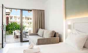 Et moderne hotelværelse med en dobbeltseng, en grå sofa og en åben glasdør, der fører til en terrasse med stole og grønt. (AI-genereret tekst)