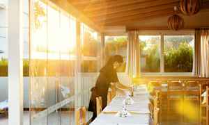 En restaurant med dækkede borde og sollys, der skinner gennem glasvæggene, potteplanter ses udenfor vinduet. (AI-genereret tekst)