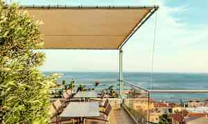 Terrasse med borde og stole, et tag af stof og udsigt over havet og huse. (AI-genereret tekst)