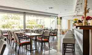 En hotelrestaurant med borde og stole samt en bardisk, store vinduer mod udendørs. (AI-genereret tekst)