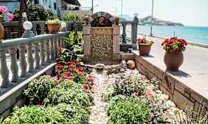 En lille fontæne omgivet af blomster og planter ved havet. (AI-genereret tekst)