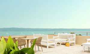 Udendørsmøbler med bord og stole i lyse farver på en terrasse med havudsigt. (AI-genereret tekst)