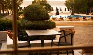 Et udendørs bord med to stole på en terrasse, med en pool og liggestole i baggrunden. (AI-genereret tekst)