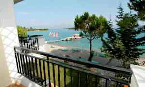 Udsigt fra en altan mod en strand med liggestole ved en klarblå havbugt. Træer og skyer i baggrunden. (AI-genereret tekst)