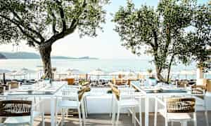 Udendørs servering med borde og stole under træer, udsigt over havet og parasoller på stranden. (AI-genereret tekst)