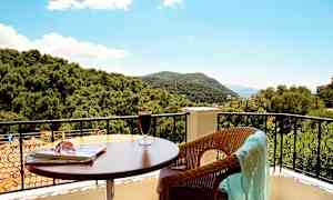 Udsigt fra en balkon med bord, drik og en stol med et klæde samt træer og bjerge i baggrunden. (AI-genereret tekst)