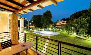 Udsigt fra en altan med bord og stole, der viser en have med oplyst pool og omkringliggende bygninger ved aftenstid. (AI-genereret tekst)