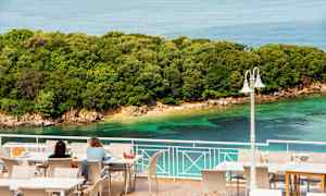 To personer sidder ved borde på en terrasse med udsigt over havet og en frodig kystlinje. (AI-genereret tekst)