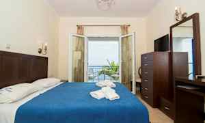 Hotelværelse med dobbeltseng, blå sengetæppe, og udsigt til havet gennem åbne glasdøre. (AI-genereret tekst)