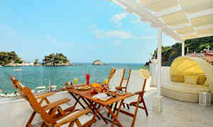 Udsigt fra en terrasse med spisebord og solseng mod havet og små øer. (AI-genereret tekst)