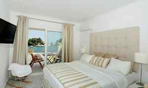 Et hotelværelse med en stor seng, beige indretning og udsigt over havet fra en balkon. (AI-genereret tekst)