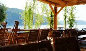 Udendørs terrasse med fletstole og borde under et trætag, omgivet af bambus og med udsigt over hav og grønne bjerge. (AI-genereret tekst)