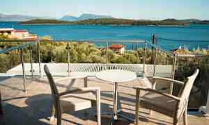 To stole og et lille bord på en terrasse med glasrækværk, udsigt til havet og skovklædte øer. (AI-genereret tekst)