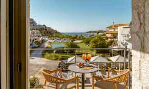 Udsigt fra en altan med to stole og et bord, der har to glas med orange drik, mod en frodig terrasse og en havn. (AI-genereret tekst)