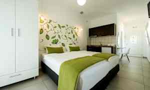 Et hotelværelse med en dobbeltseng dækket med hvidt sengetøj og grønne tæpper, et klædeskab, et tekøkken og en dør i baggrunden. (AI-genereret tekst)