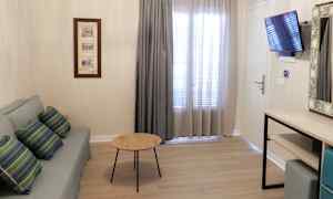 En stue med sofa, bord, gardiner foran vinduet, en vægmonteret tv og en kommode. (AI-genereret tekst)