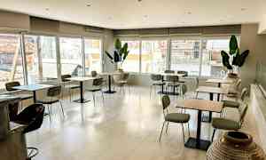 En lys café med borde og stole samt store vinduer, der lukker naturligt lys ind. Flere potteplanter pynter rummet. (AI-genereret tekst)