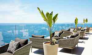 Loungeområde med rattanstole, store potteplanter og udsigt over havet. (AI-genereret tekst)