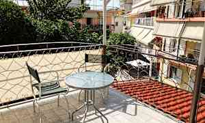 Altan med glasbord og to metalstole, med udsigt til grønne områder og omkringliggende bygninger. (AI-genereret tekst)