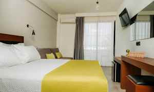 Et hotelværelse med en dobbeltseng med hvidt sengetøj og gult sengetæppe, en sofa med puder, et skrivebord med spejl og et TV på væggen. Altandøre med lyse gardiner. (AI-genereret tekst)