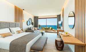 Et stilfuldt hotelværelse med en dobbeltseng, sofa og et skrivebord. Store glasdøre åbner ud til en altan med udsigt over havet. (AI-genereret tekst)