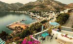 Udsigt fra terrassen over en havn med både, omkringliggende bygninger og bjerge i baggrunden. (AI-genereret tekst)