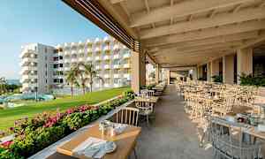 En udendørs restaurant med dækkede borde, grønne områder og en flerfarvet bygning i baggrunden. (AI-genereret tekst)