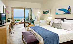 Et hotelværelse med dobbeltseng, sofa og stor glasdør, der fører til en terrasse med udsigt over havet. (AI-genereret tekst)