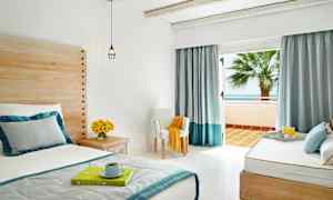 Et lyst og luftigt hotelværelse med seng, træpaneler, blomster på et sidebord og åbne glasdøre, der fører ud til en balkon med havudsigt. (AI-genereret tekst)