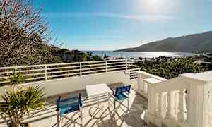 Udsigt fra en terrasse med bord og stole, med havet og bjerge i baggrunden. (AI-genereret tekst)